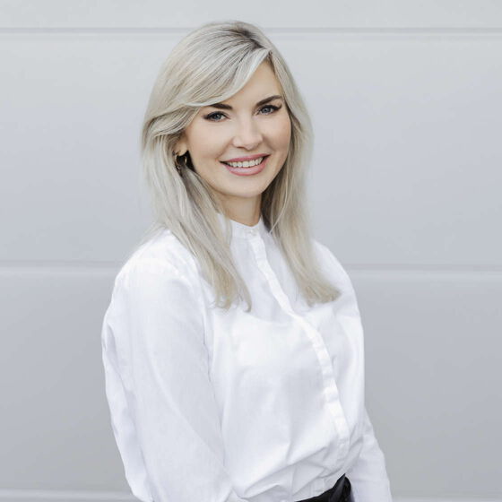 Fotografie von Angelina Diab, Geschäftsführerin der Reinigungsfirma PLAZA Cleaning für Gebäudereinigung und Gewerbereinigung wie Büroreinigung, Praxisreinigung, Kita-Reinigung, Schulreinigung. Lächelnde gepflegte Frau trägt weiße Bluse und schwarzen Rock.